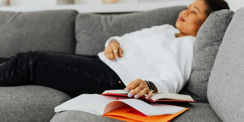 Persona descansando en el sofá junto a un libro, imagen asociada a la importancia de la siesta y el descanso durante el día